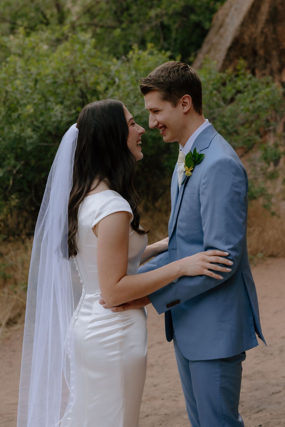 Bride and groom laughing together outdoors near red rock formations in desert landscape.