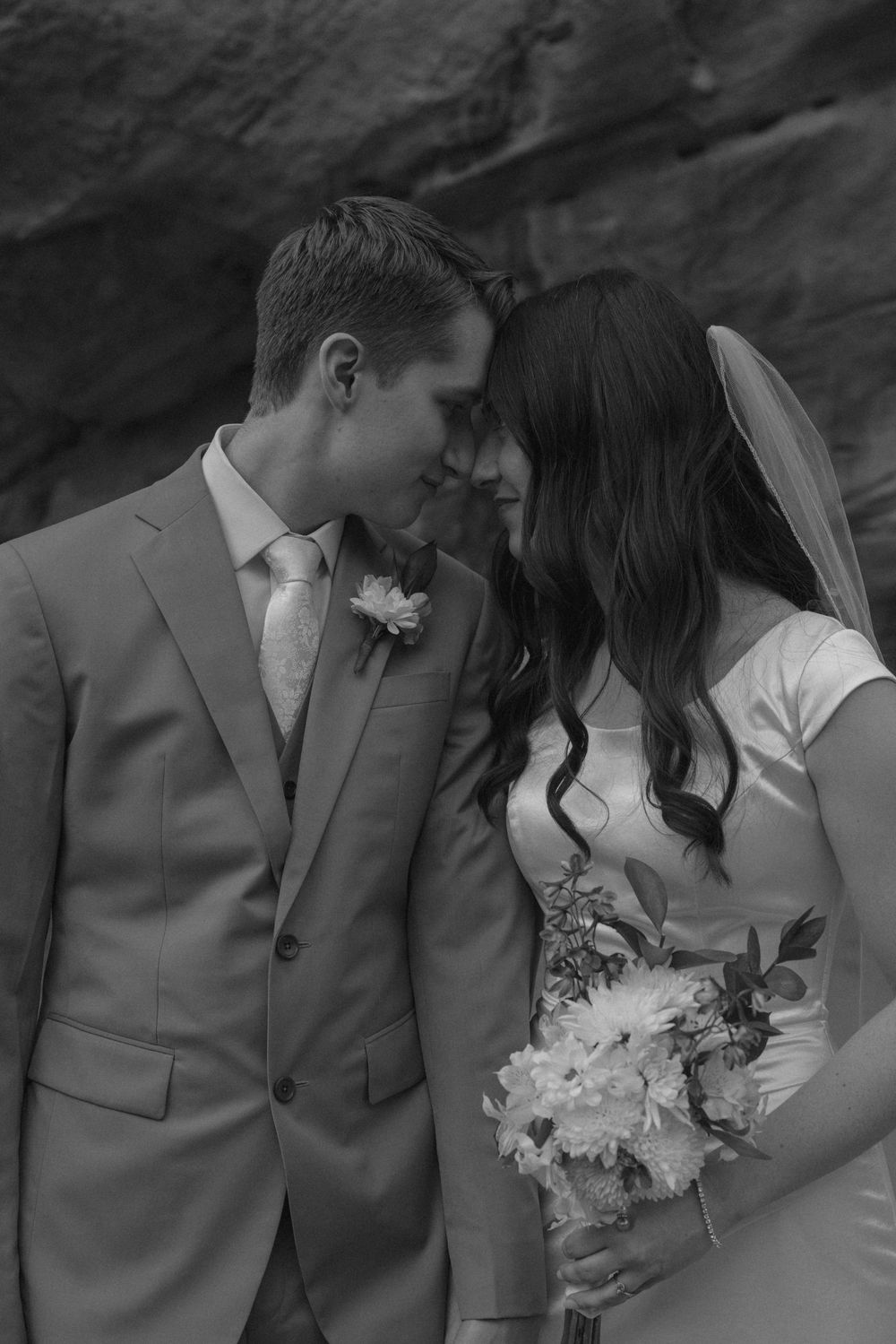 Black and white wedding photo of bride and groom touching foreheads, bride holding bouquet, rocky background.