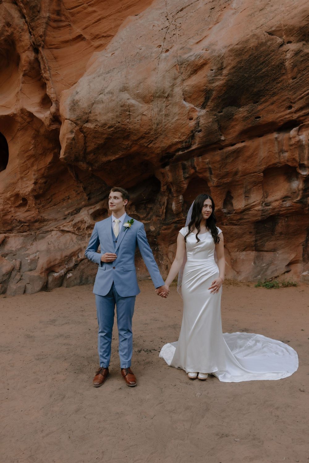 Bride and groom hold hands in white dress and blue suit before red rock canyon walls.