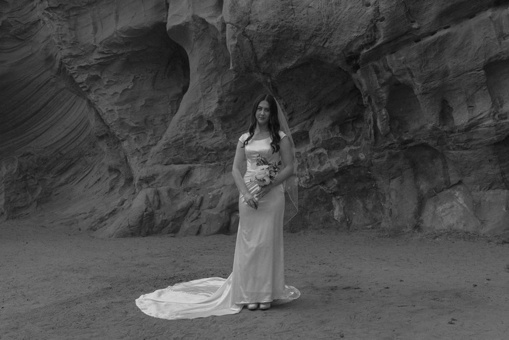 Black and white photo of bride in white gown with long train standing alone before red rock canyon walls.
