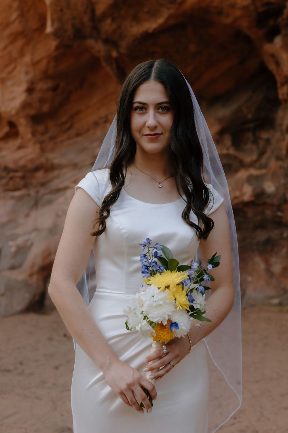 Bride in white satin gown and veil holds colorful bouquet before red rock canyon walls.