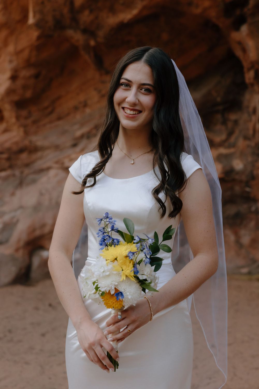 Smiling bride in white gown holds blue and yellow bouquet against red rock backdrop.