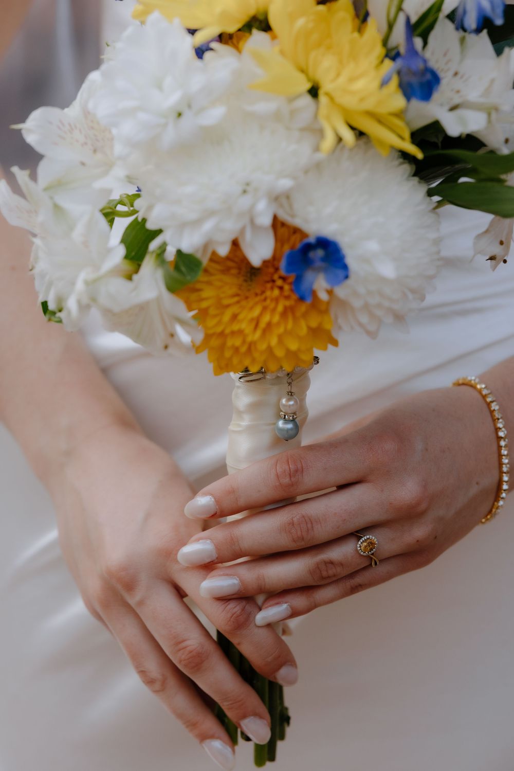 Bride holding a wedding bouquet of white, yellow, and blue flowers with manicured nails, gold ring, and bracelet.