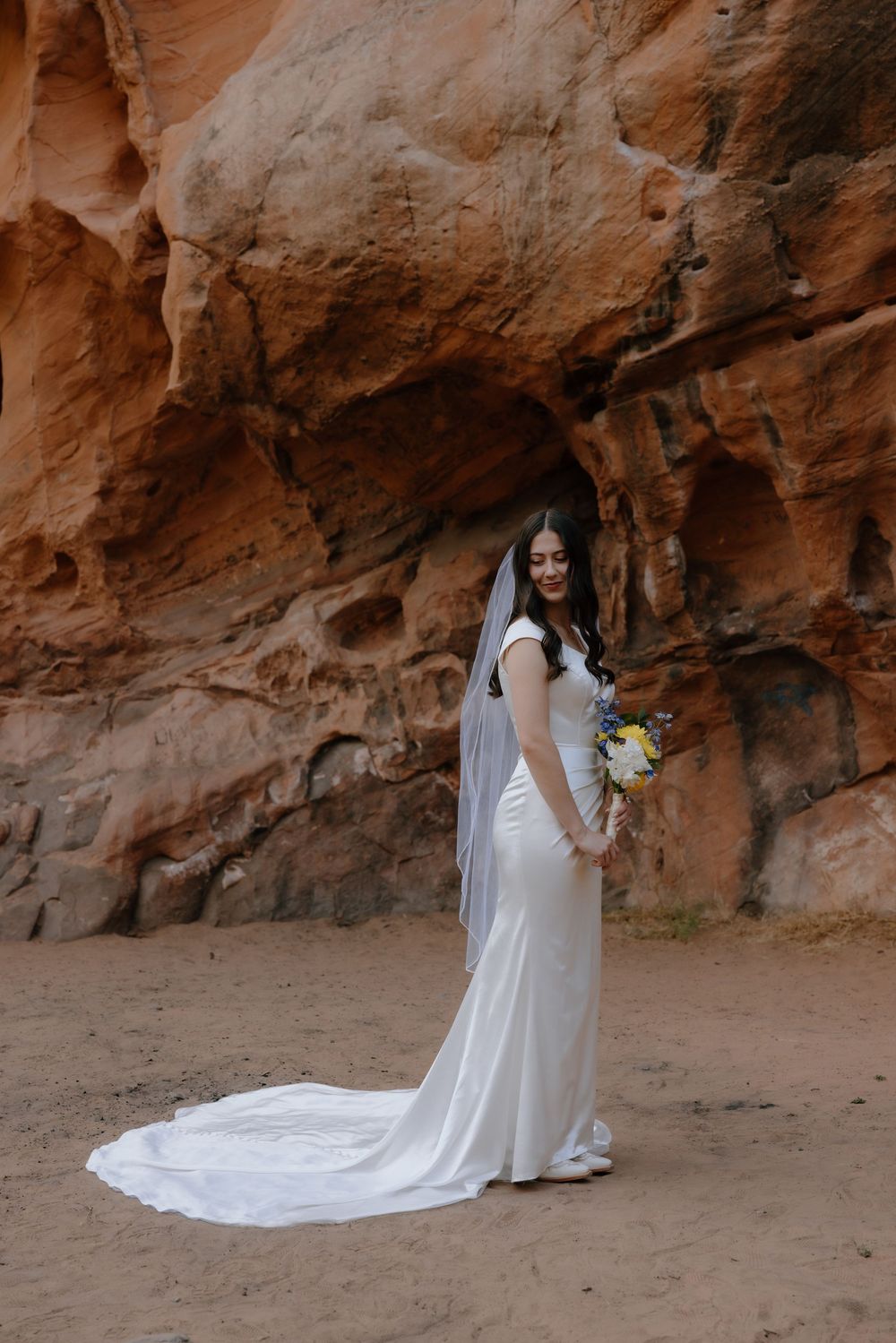 Elegant bride in fitted white dress with long train poses against red sandstone rock formation.