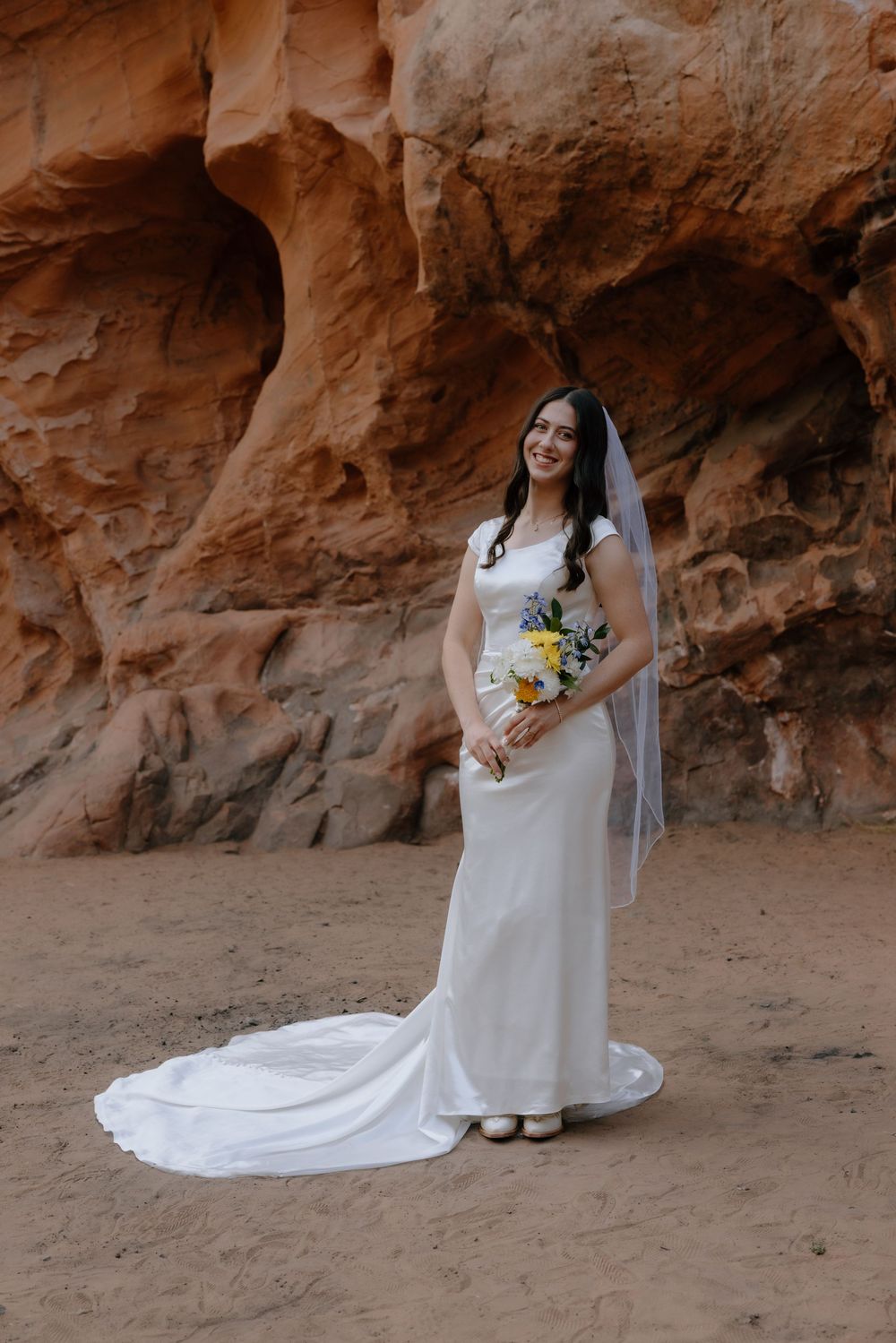 Smiling bride in white gown and veil holds colorful bouquet before red rock canyon walls.