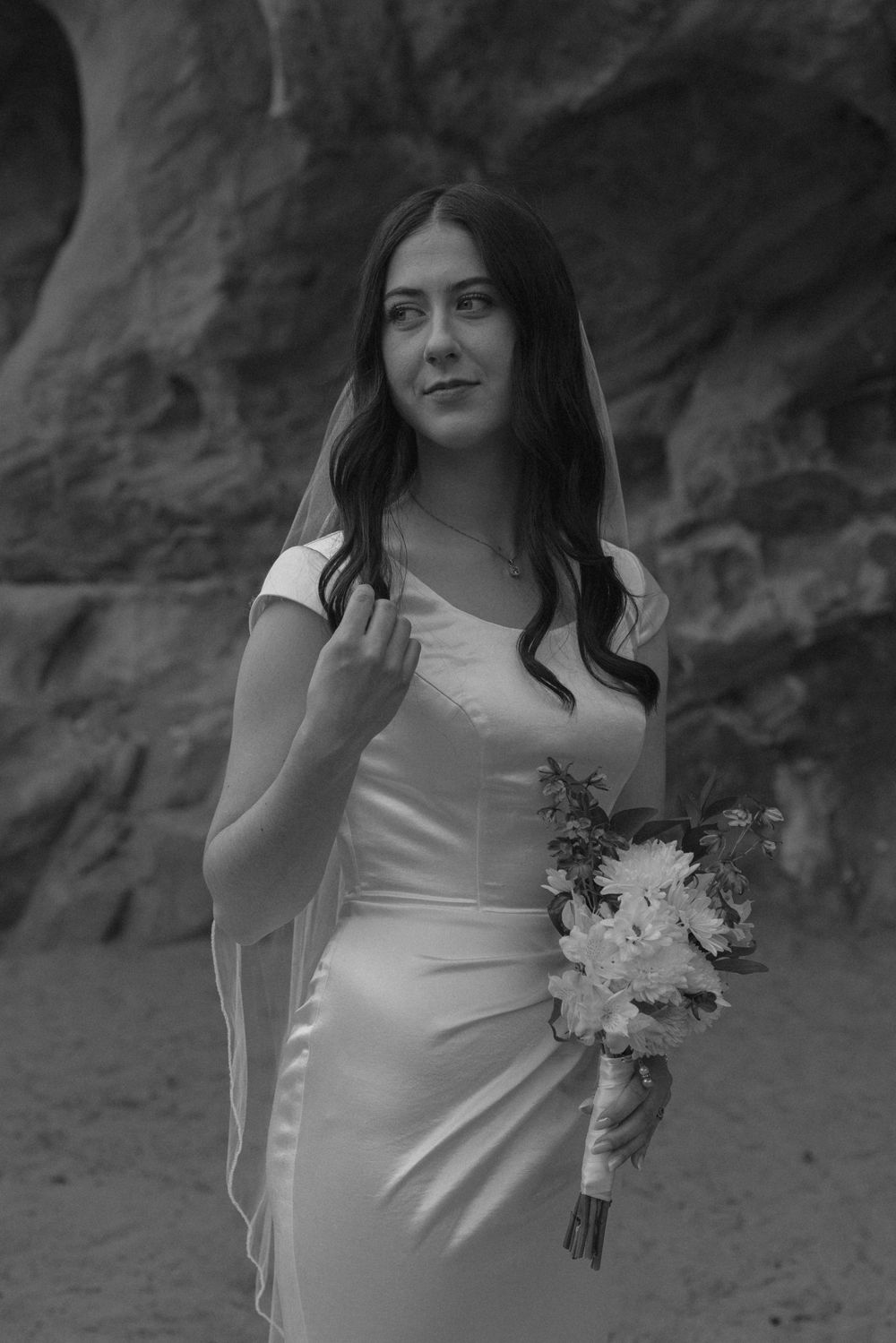 Black and white portrait of bride in satin gown holding bouquet before canyon rock walls.