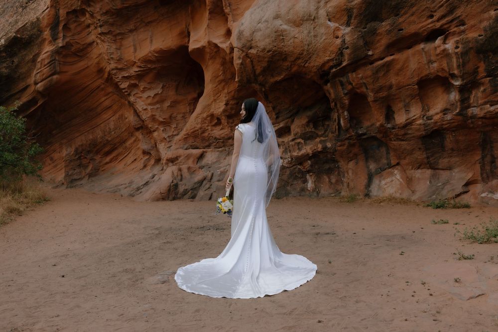 Bride in white wedding gown and veil stands before red rock canyon walls holding a bouquet.