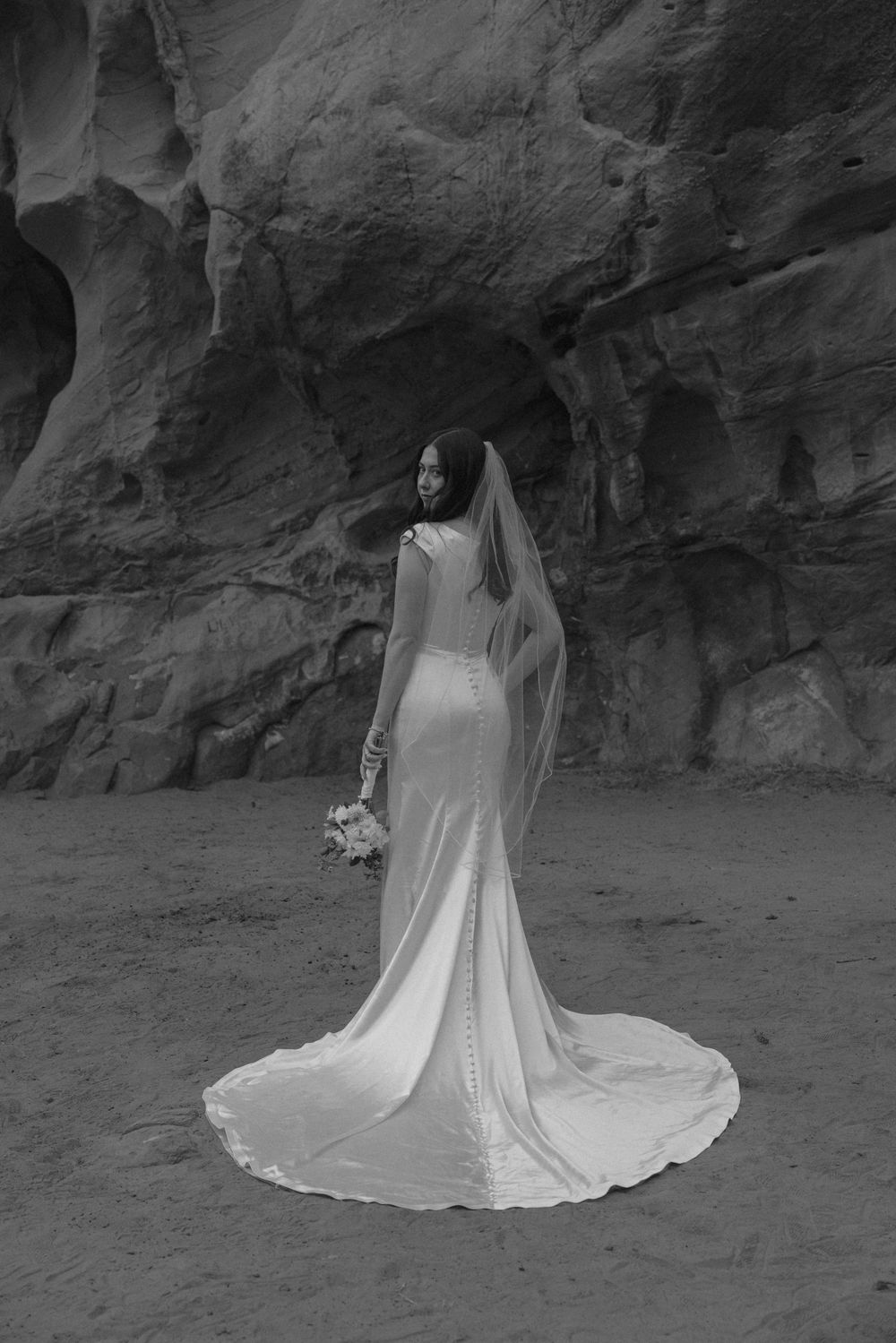 Black and white photo of bride in fitted gown and veil standing before dramatic rock walls.