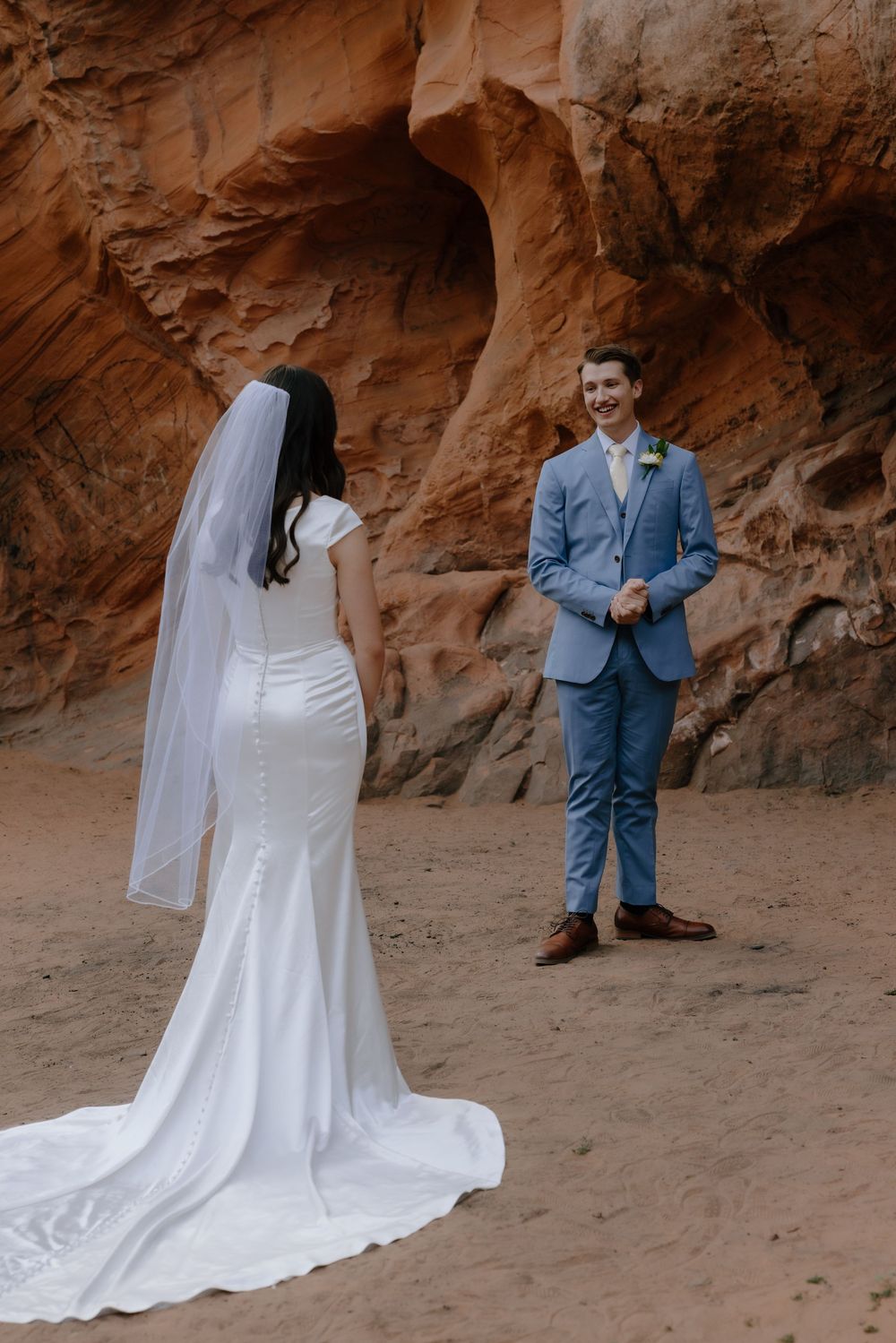 Bride in white gown and veil faces groom in blue suit during outdoor desert elopement ceremony near red rock formations.