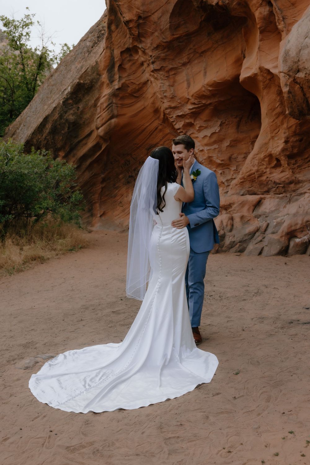 Bride and groom laugh together in red rock canyon, her mermaid gown trailing on sand.
