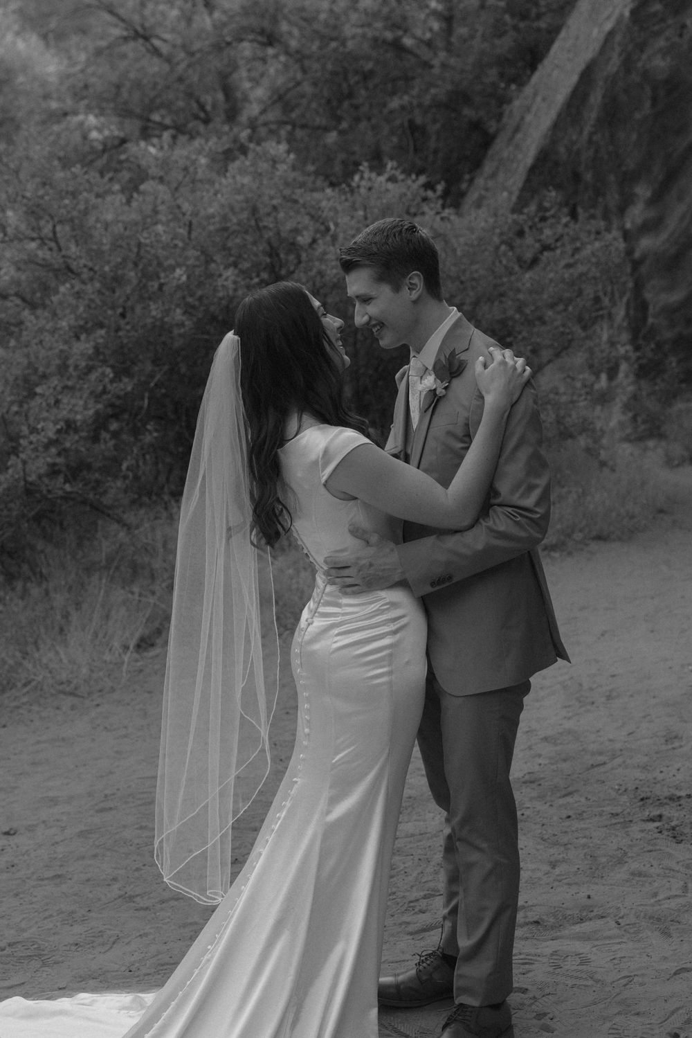 Black and white photo of a bride and groom embracing outdoors, with the bride in a fitted gown and veil.