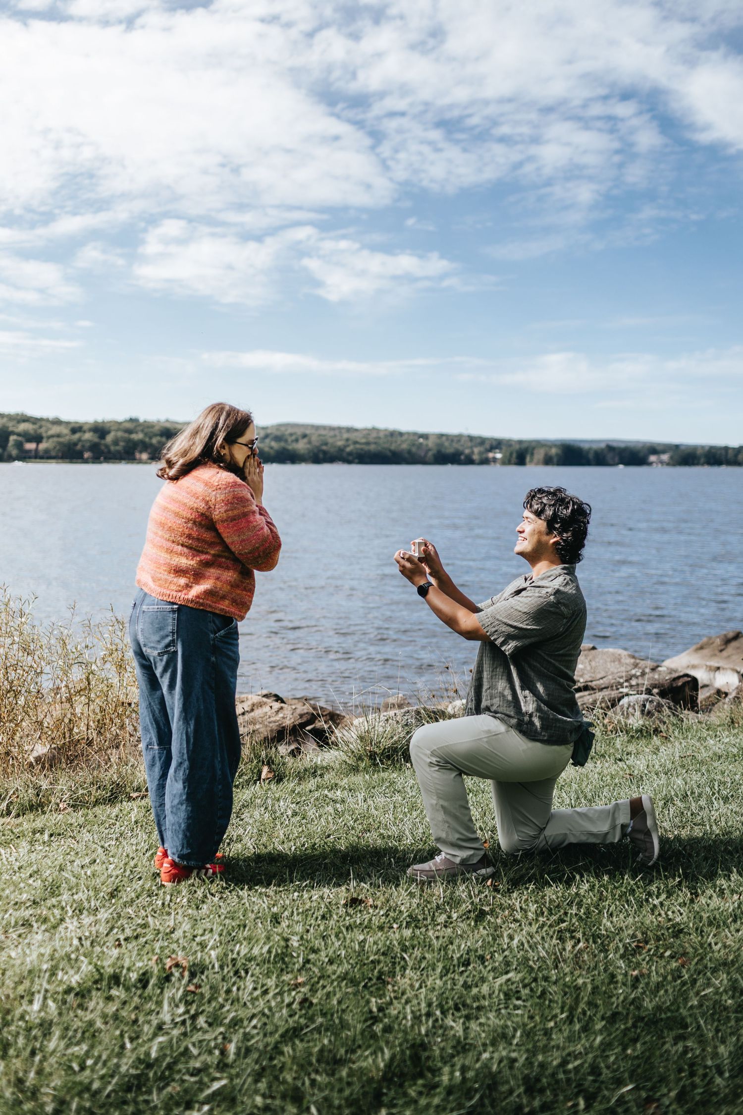 Man proposes on one knee to woman by a scenic lakeside on a sunny day with blue skies and green hills.
