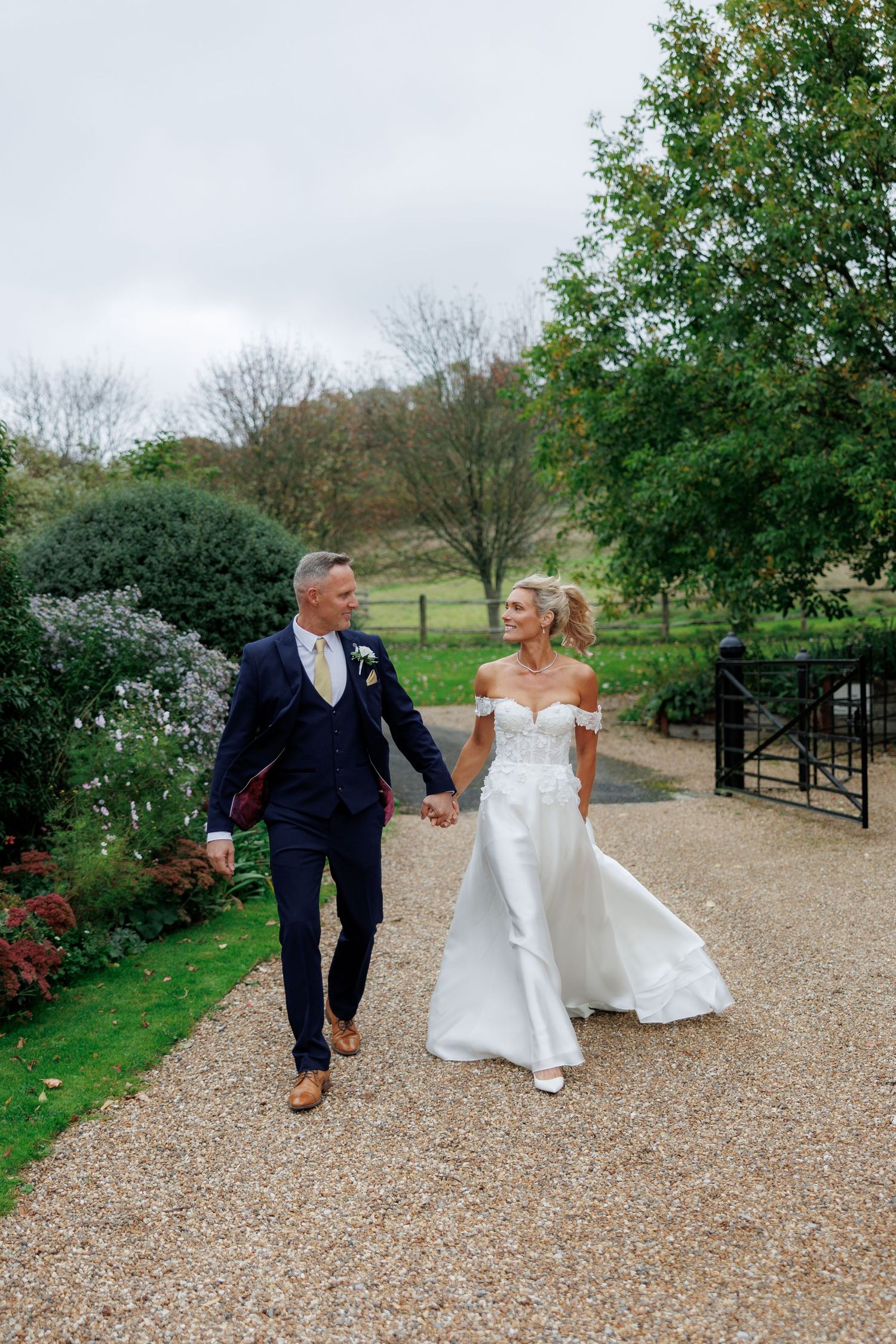 Bride in an off-shoulder white wedding gown and groom in a navy suit hold hands walking along a gravel path beside a garden.