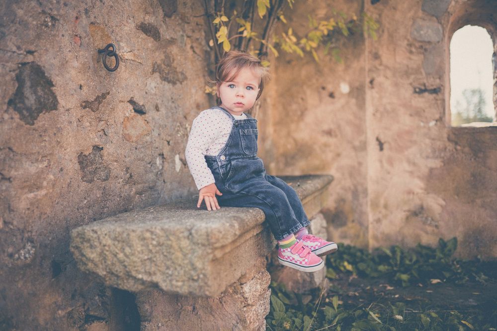 Toddler in denim overalls and pink sneakers sits on a stone ledge inside rustic ruins with autumn leaves on the ground.