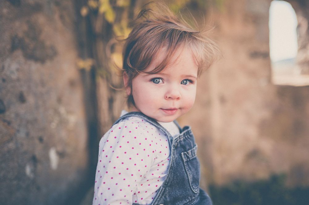 Toddler with light brown hair, wearing denim overalls over a polka dot shirt, smiles softly while looking over the shoulder outdoors.