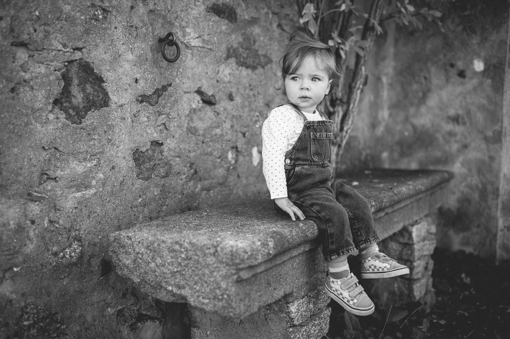 Black and white photo of a toddler in denim overalls and sneakers sitting on a weathered stone bench against an old rustic wall.