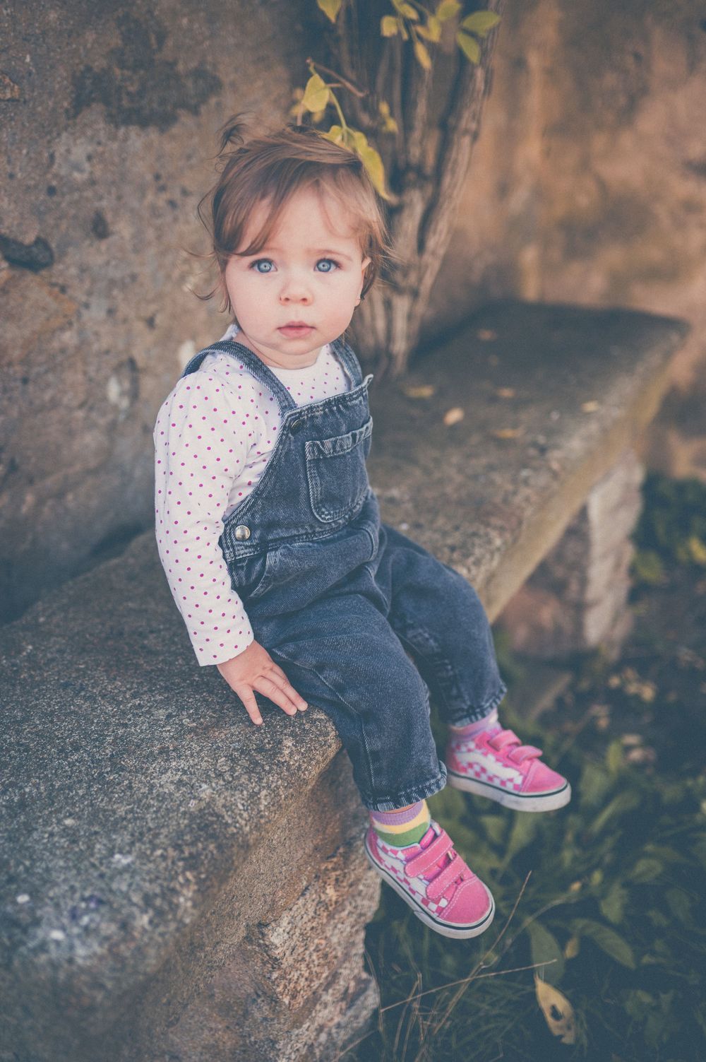 Toddler in denim overalls and pink sneakers sits on a weathered stone bench beside autumn leaves, looking at the camera.