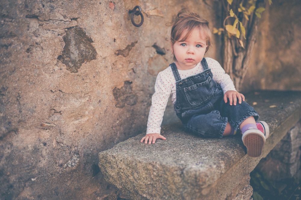 Toddler in denim overalls and a white long-sleeve shirt sits on a rough stone ledge beside a weathered rustic wall.