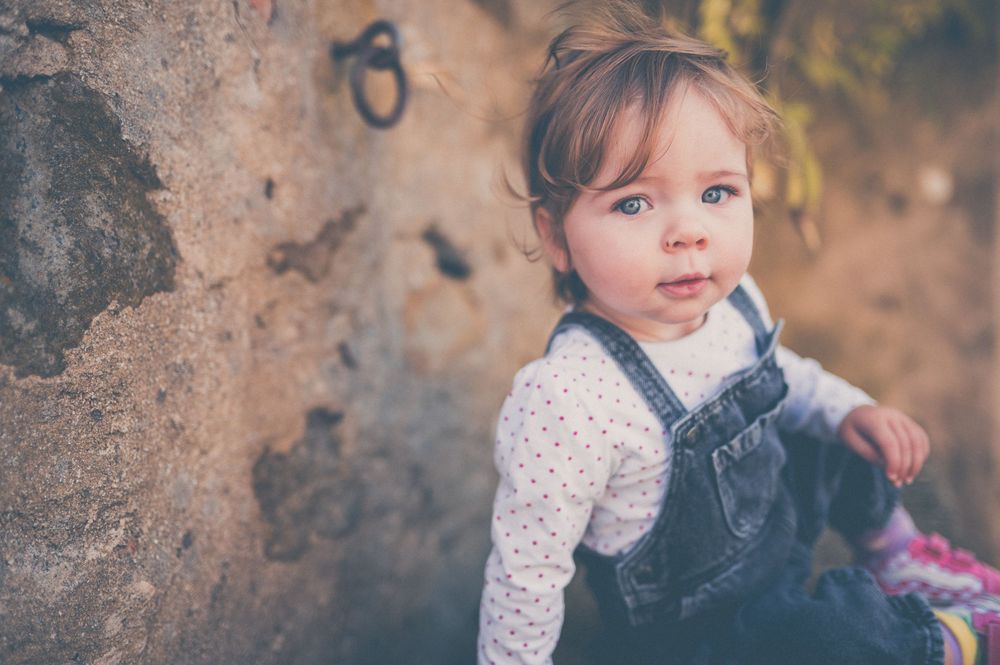 Curious toddler with blue eyes wearing a denim overall dress and polka dot shirt sits beside a rustic stone wall.