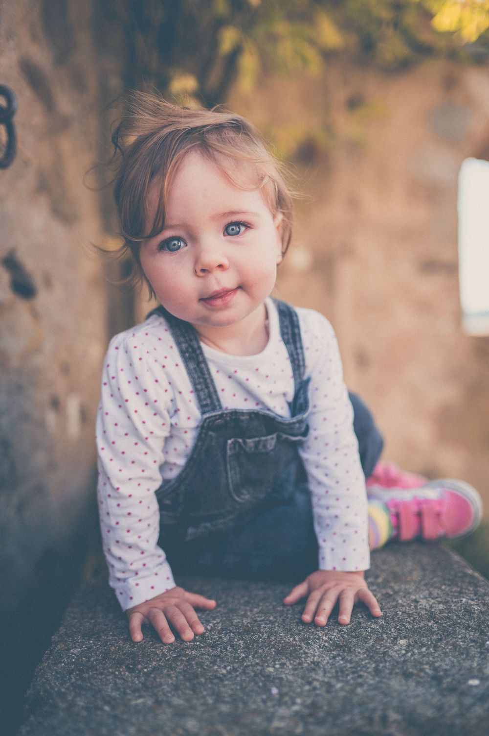 Smiling blue-eyed toddler in denim overalls and pink shoes crawling on a stone ledge outdoors in soft golden light.