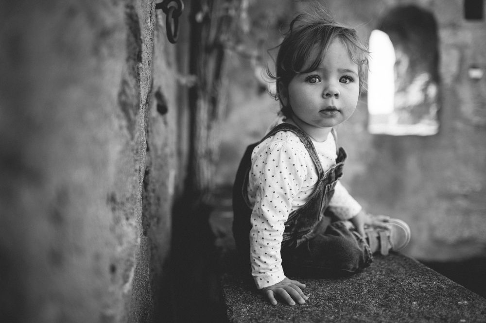 Black and white portrait of a toddler in denim overalls sitting on a stone ledge and gazing at the camera.