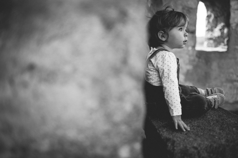 Black and white photo of a toddler in overalls and a patterned shirt sitting on a stone ledge, gazing out a window.