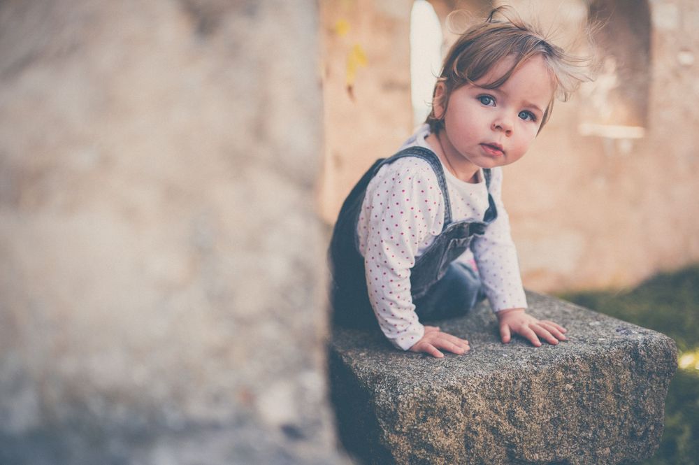 Toddler in a gray pinafore and white polka dot shirt leans on a mossy stone ledge, gazing at the camera in a soft-focus outdoor setting.