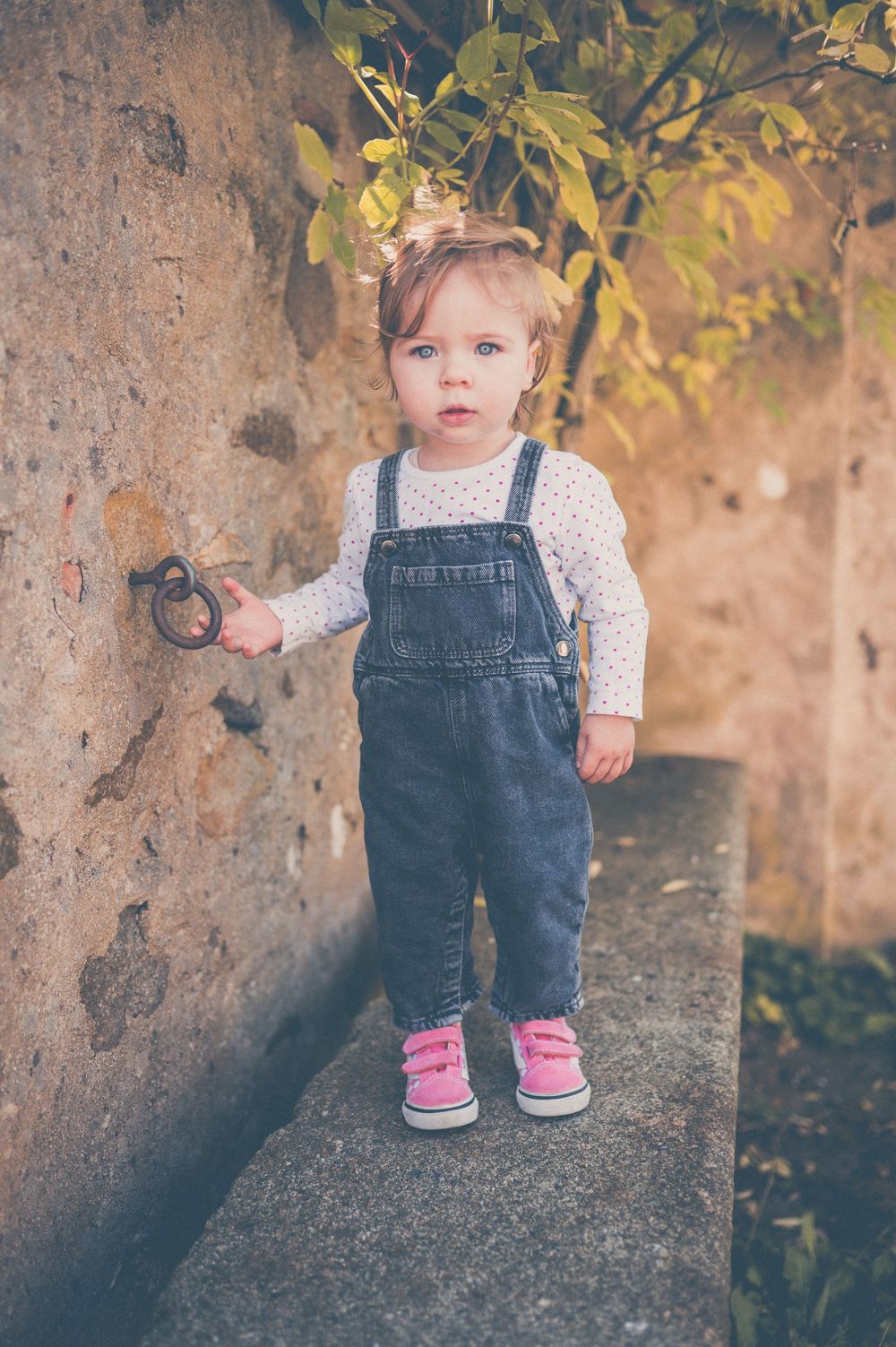 Toddler in denim overalls and pink sneakers stands on a stone ledge holding an iron ring on a rustic wall.