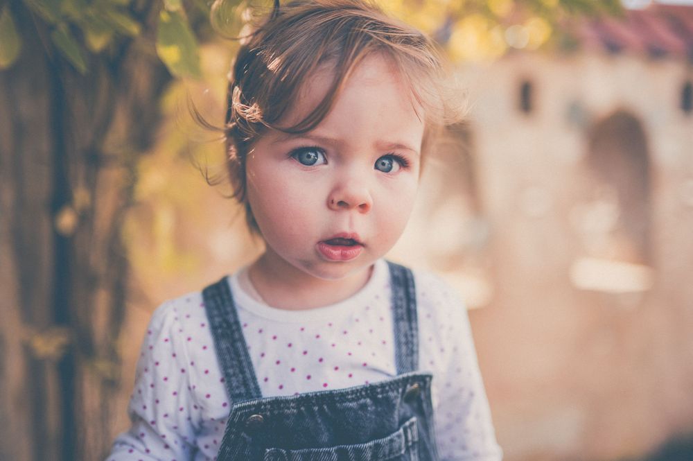 Curious toddler with blue eyes and a small hair bow wears denim overalls outdoors against a softly blurred warm background.