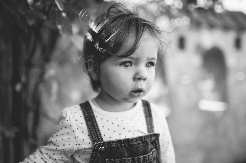 Black and white portrait of a curious toddler in denim overalls and a polka dot shirt gazing thoughtfully outdoors.