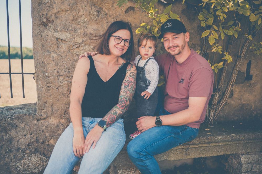 Smiling family of three, a woman with glasses and tattoos, a man in a cap, and a toddler, posing on a stone bench outdoors.