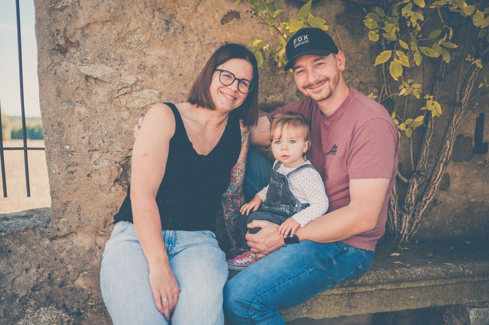 Smiling mother and father in a cap sit on a stone bench holding their toddler in denim overalls during a family photo session.