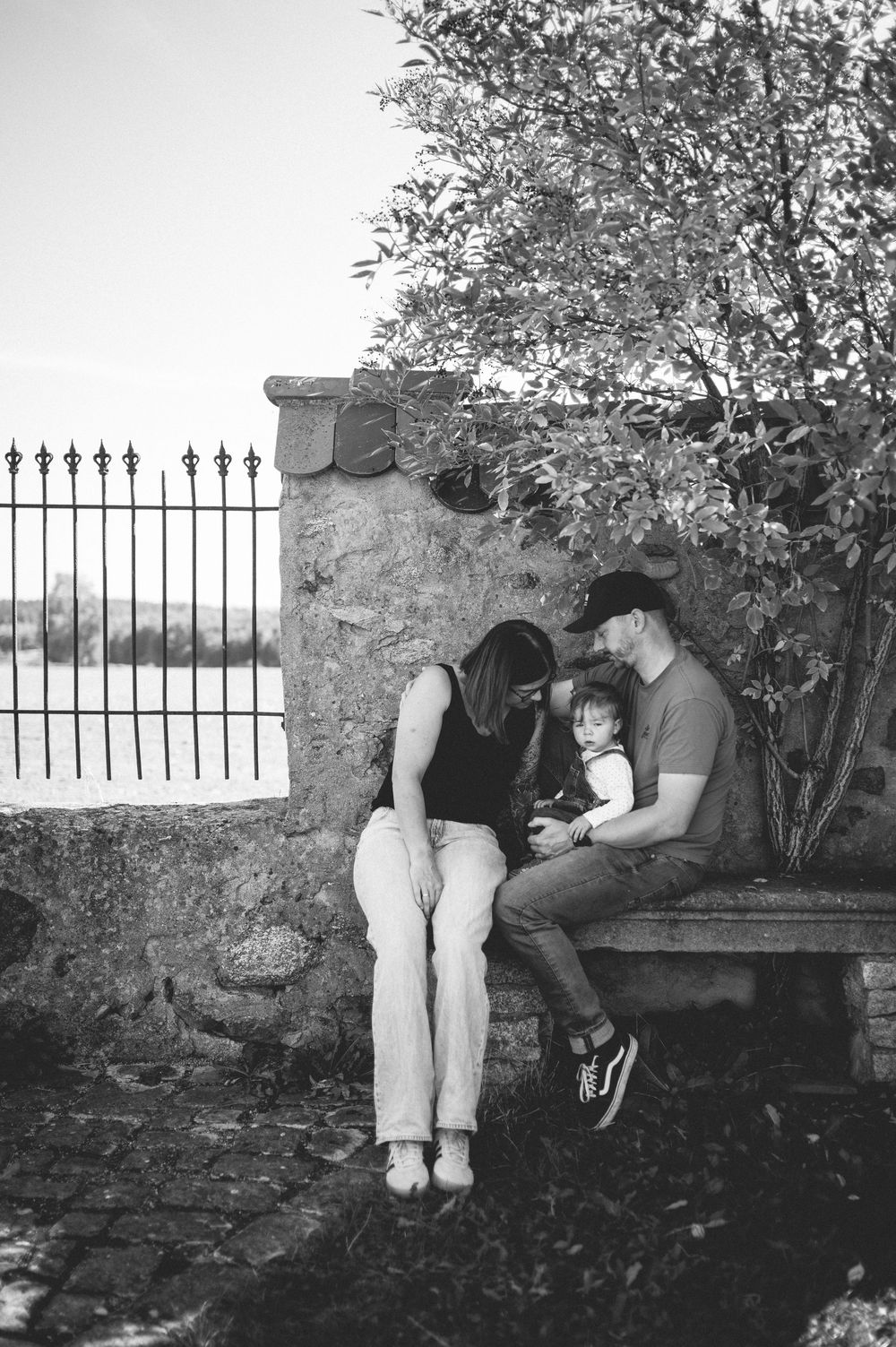 Black and white photo of a mother and father in caps sitting on a stone bench with their baby beside an iron fence and tree.