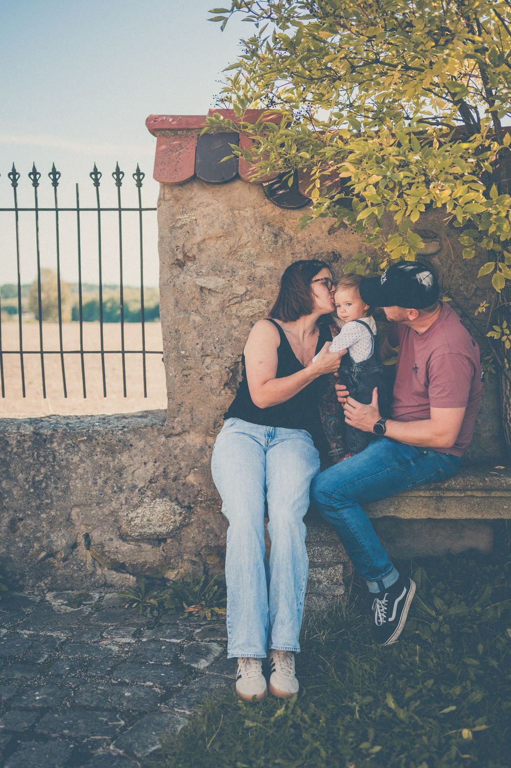 A couple sits on a stone bench beneath a leafy tree, kissing their baby between them, with an iron fence behind.
