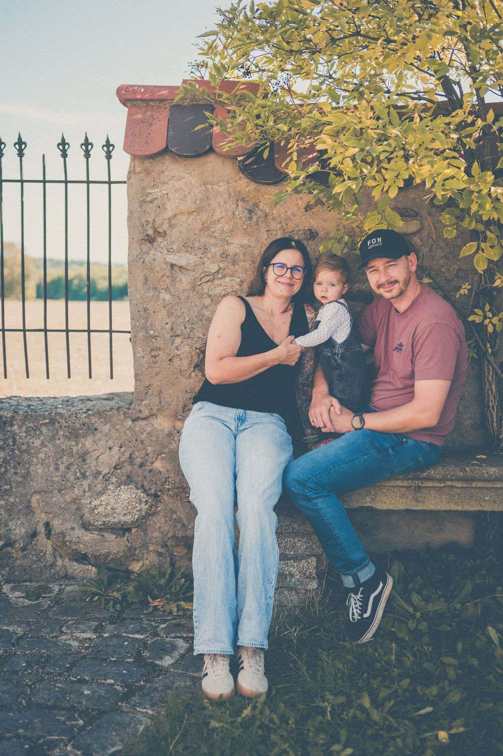 A smiling family of three sits together on a rustic stone bench near an iron fence, with a baby held in the mother's lap.