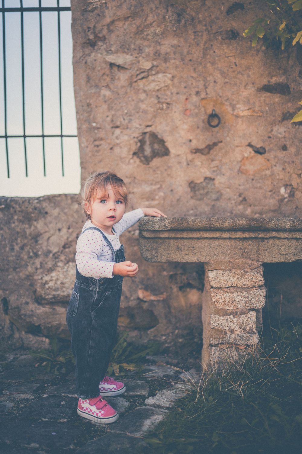 Toddler in denim overalls and pink sneakers reaches toward a weathered stone bench beside an old rustic stone wall.