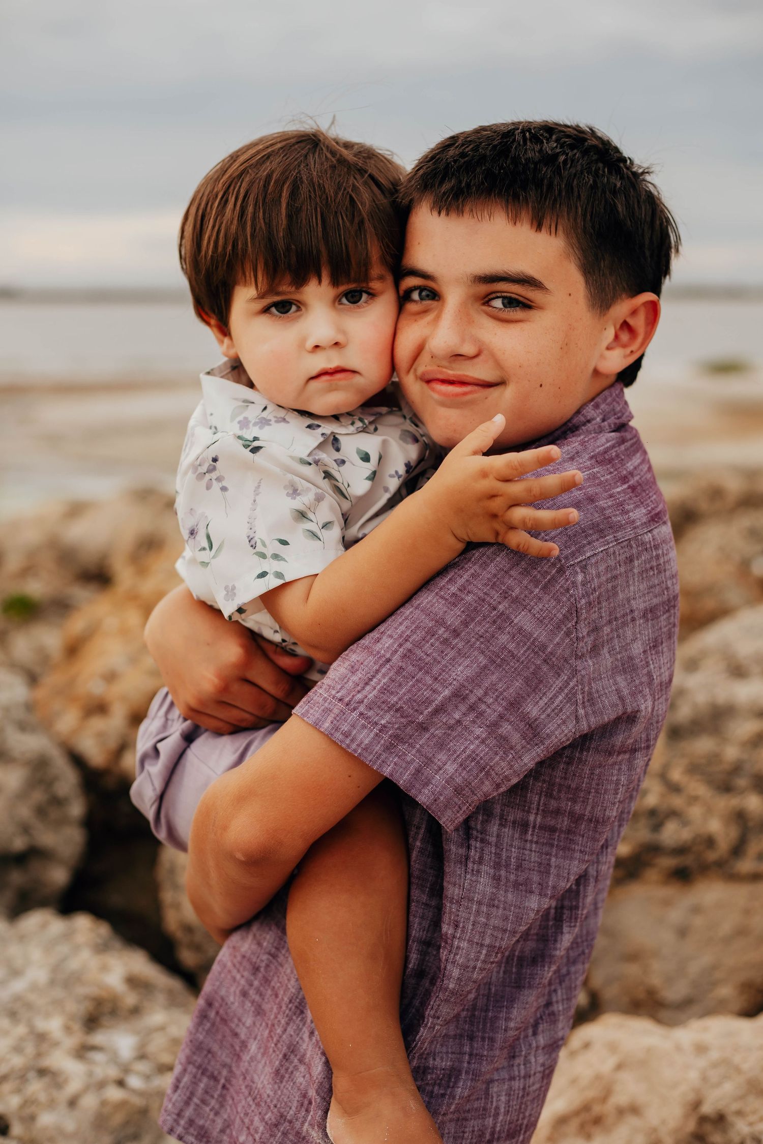 Smiling older boy in a purple shirt holds a toddler in a floral shirt in a warm embrace on a rocky beach at sunset.