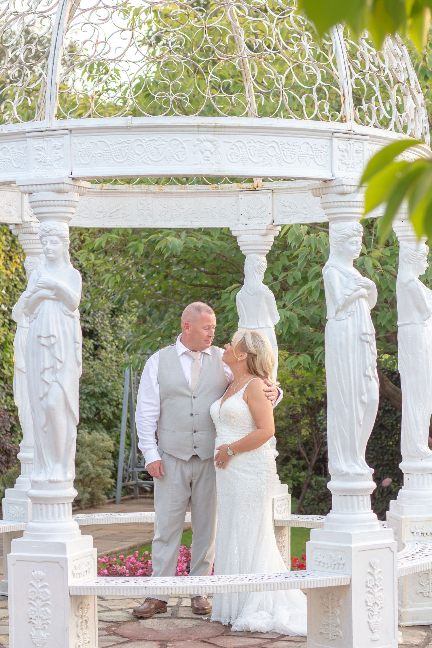 Bride in a white gown and groom in a gray vest and trousers embrace inside an ornate white gazebo with classical statue columns.