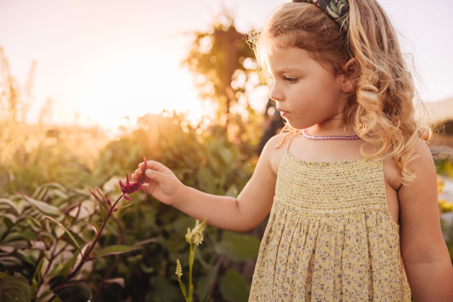 Young girl in a yellow floral sundress gently touching a pink flower in a sunlit garden at golden hour.
