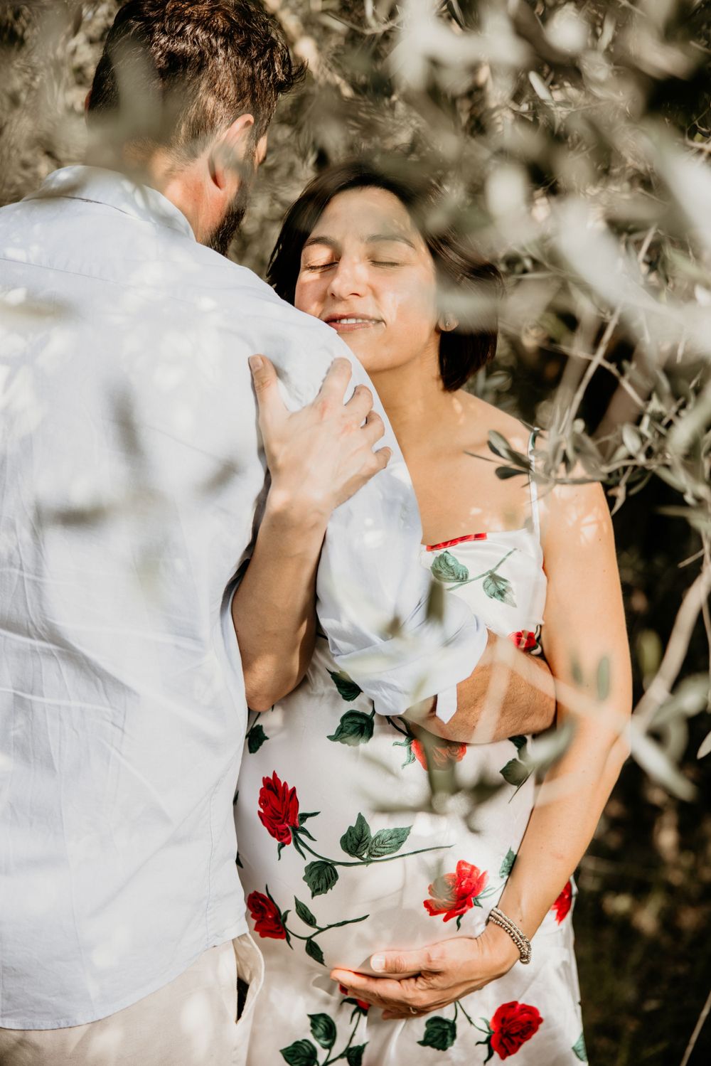 Pregnant woman in floral dress embraces partner among olive tree branches in a tender maternity photo.