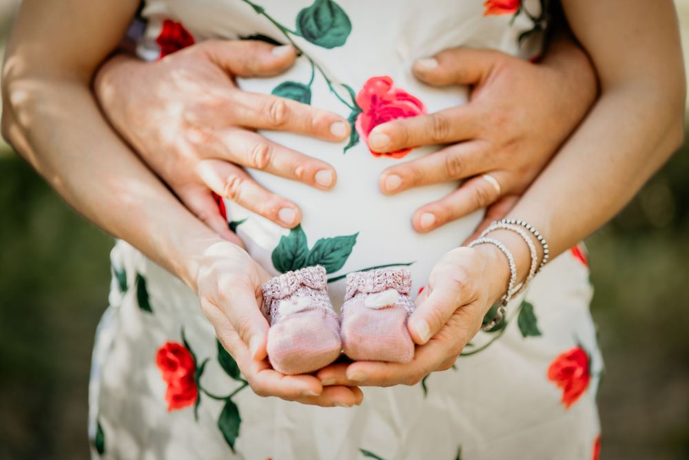 Couple holding tiny pink baby shoes over pregnant belly in floral dress during maternity session.