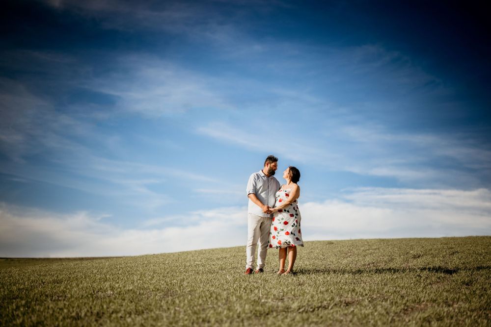 Expectant couple standing together in open green field under dramatic blue cloudy sky.
