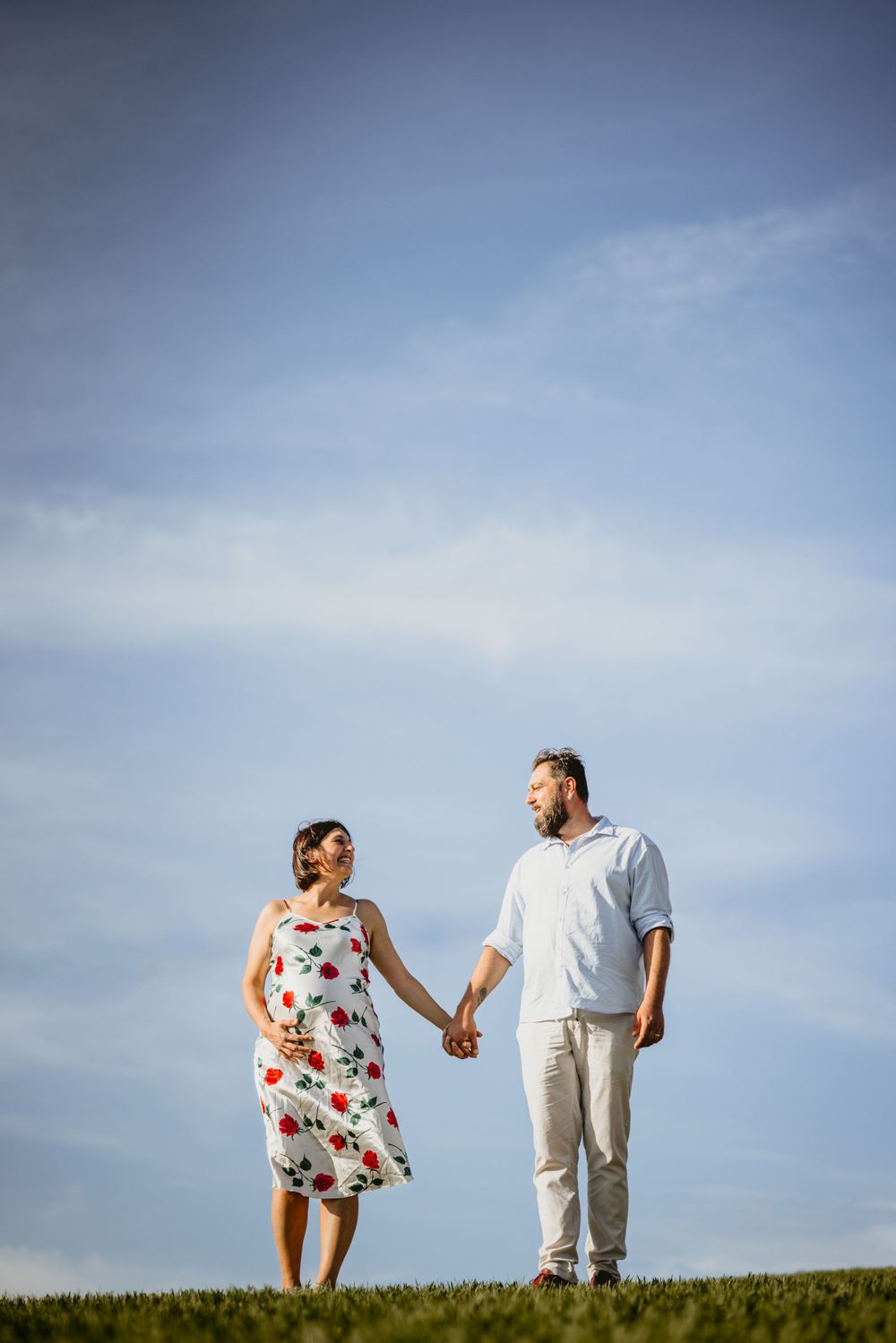 Pregnant couple holding hands walking on green grass field under blue sky.