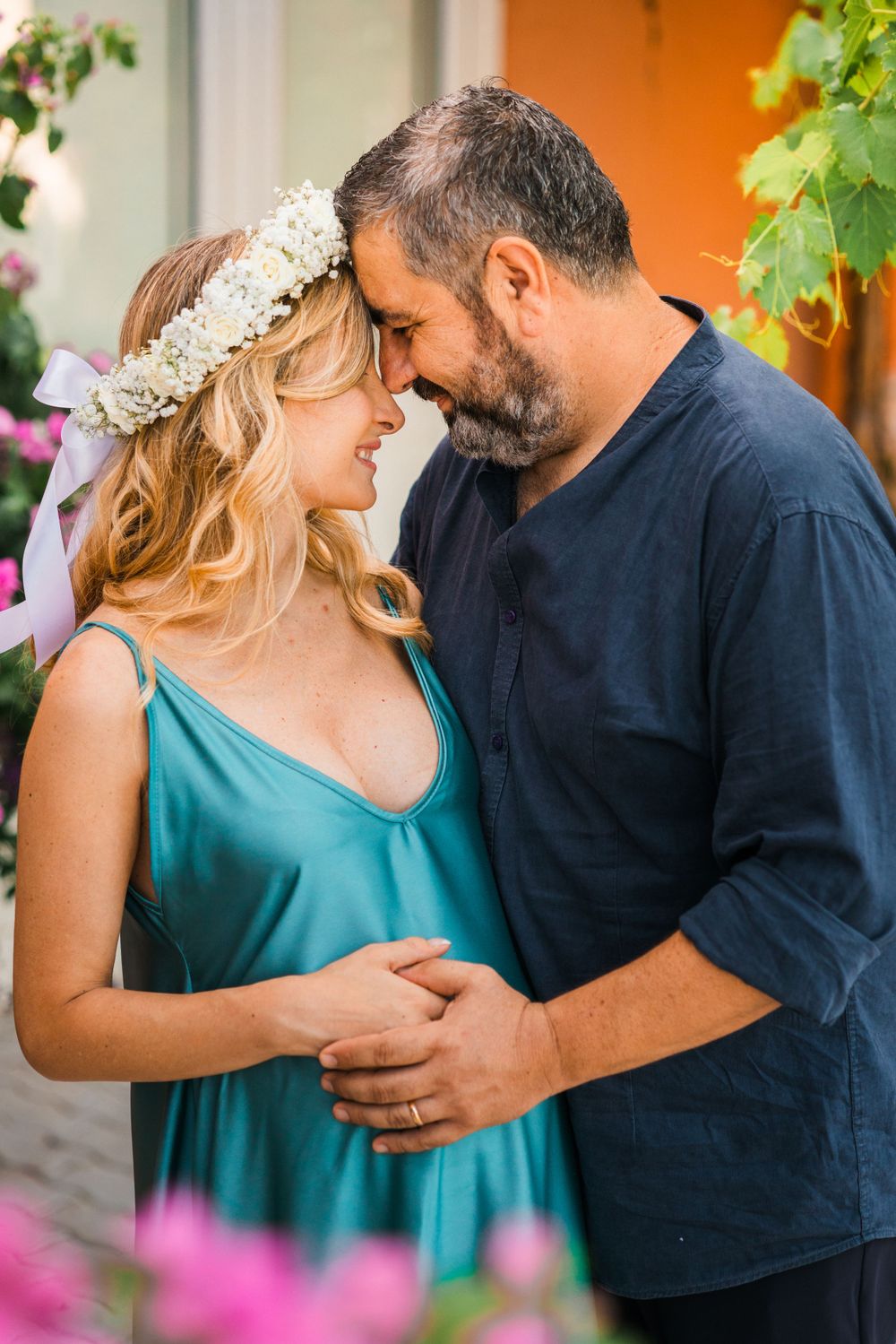 Pregnant woman in teal dress with flower crown shares intimate moment with bearded partner.