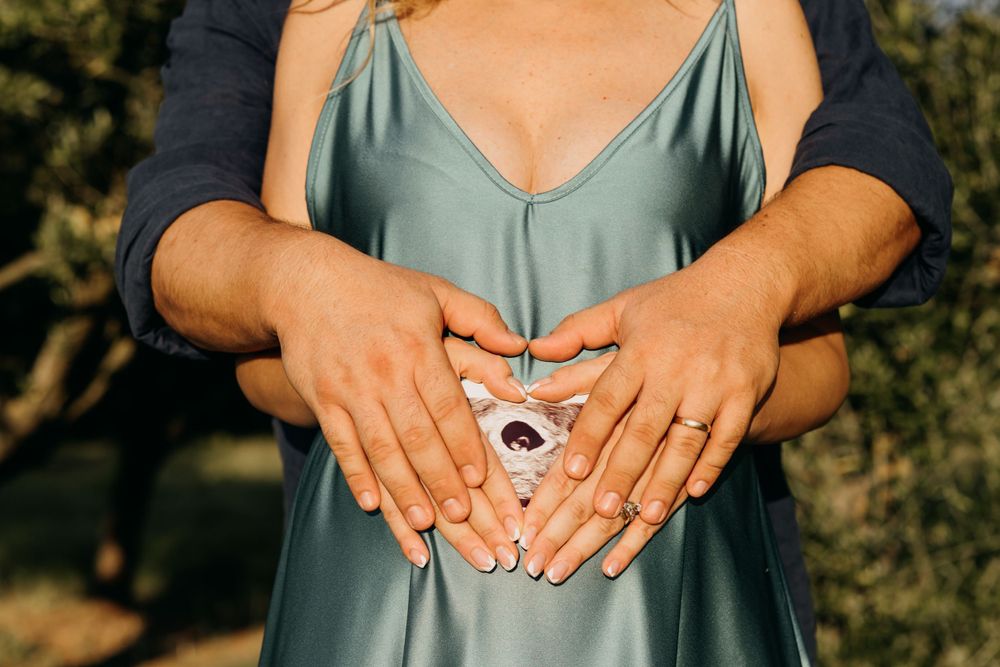 Couple forming heart shape with hands over pregnant belly in sage green maternity dress.