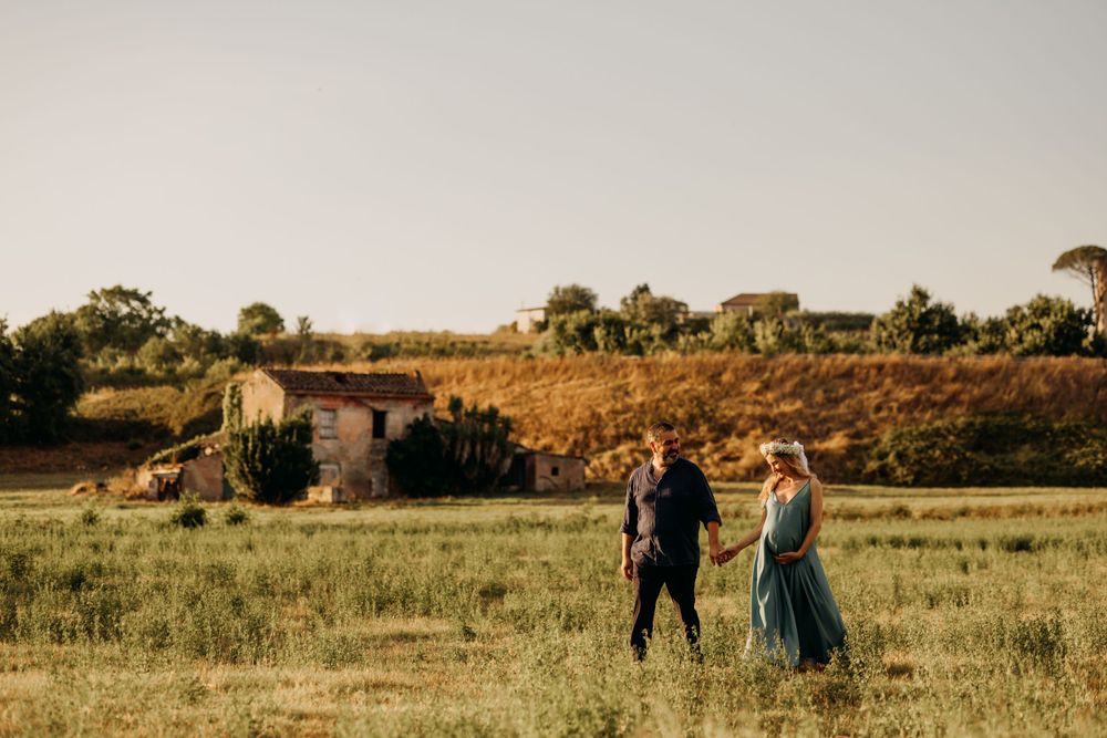 Couple walking through a sunlit green field with rustic farmhouse and golden hills in the background.