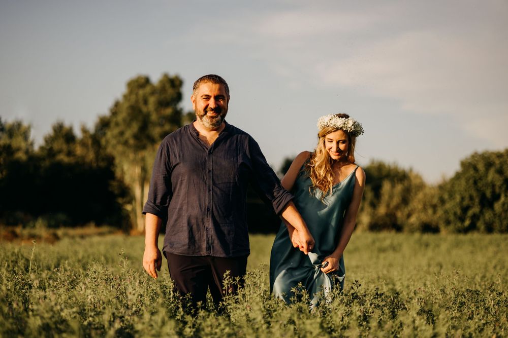 Smiling couple holding hands in a lush green field at golden hour, woman wearing floral crown.