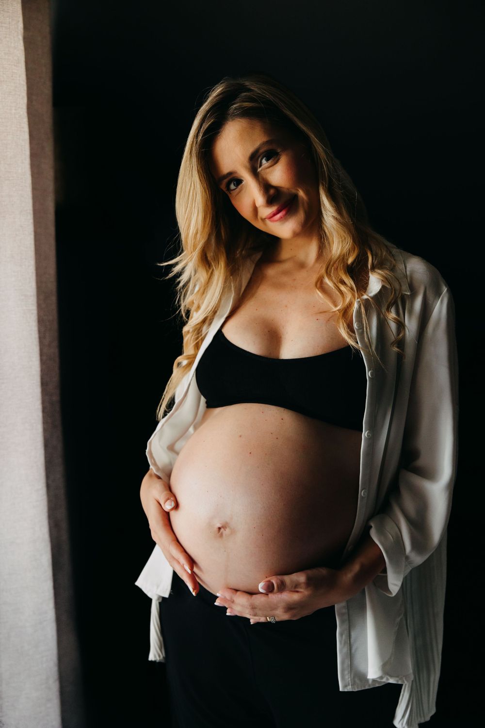 Pregnant blonde woman in black bralette and white shirt cradling her baby bump against dark background.