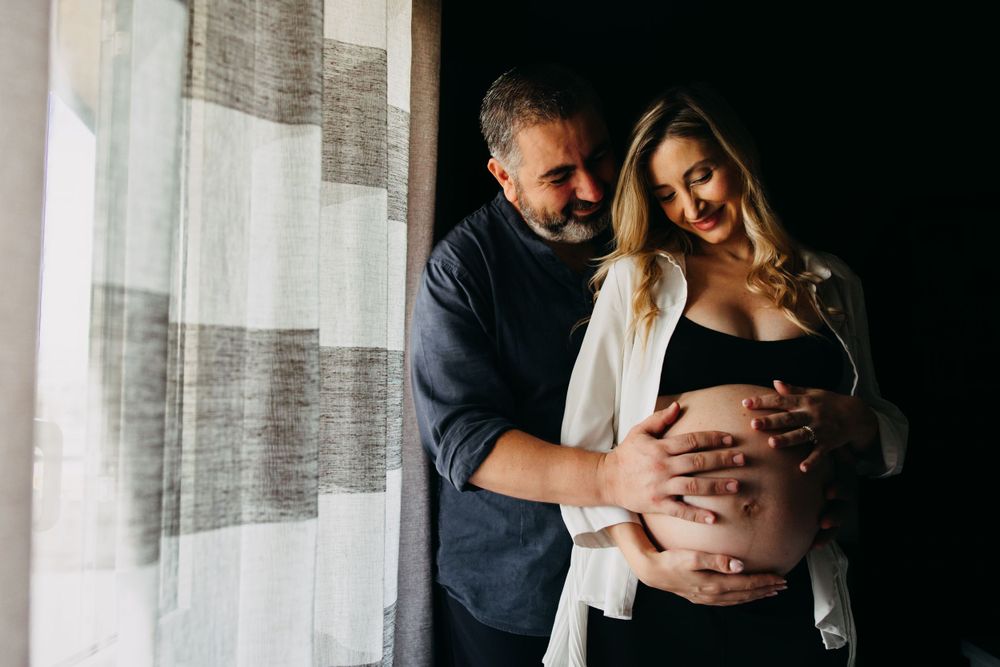 Loving couple embracing pregnant belly near a window with soft natural light indoors.