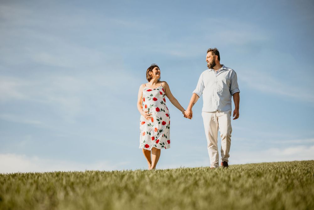 Happy pregnant couple holding hands walking barefoot through green meadow under blue sky.